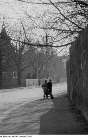 Zwei Kinder mit Kinderwagen auf dem Bürgersteig einer leeren Straße
