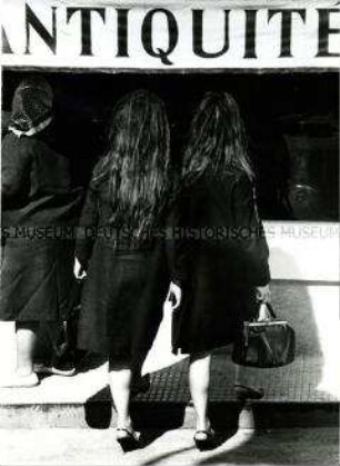 Rückenansicht zweier Frauen mit langen Haaren, in dunklen Mänteln, die nebeneinander eine Stufe hochsteigen