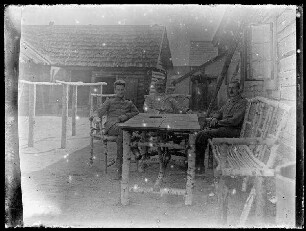 Unbekannte Ortschaft [Litauen/Weißrussland/Ukraine?]: 3 Offiziere im Freien vor einem Holzhaus auf Stühlen und einer Bank an einem Tisch, alles aus Birkenholz, im Hintergrund ein niedriges landwirtschaftliches Holzhaus mit Schindeldach