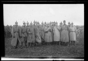 Nicht identifizierbare Gegend in Nordfrankreich oder Belgien: 17 Offiziere mit Pickelhaube und Mantel und einer mit Tschapka auf einem Feld, im Hintergrund Soldaten mit Pickelhaube und zusammengestellte Gewehre (Gruppenbild, Distanzaufnahme)