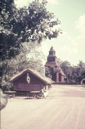 Reisefotos. Junge Frau in Tracht vor einer Imbißbude. Blick zu einer Holzkirche (vielleicht in Skandinavien)