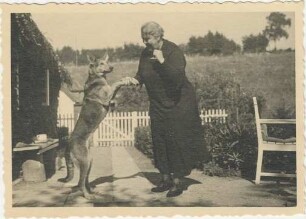 unbekannt: ältere Frau, daneben Schäferhund gibt Pfote: Rohlfshagen, Kupfermühle: Schäferkate, vor Traufseite: Plattenweg, rechts Gartenstuhl, hinten Zaun, Gartentor, dahinter Atelier, Schuppen: im Hintergrund Wiese an Abhang