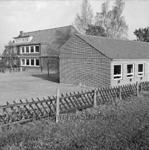 Volksschule: Schultwiete: vorn Rasen mit Jägerzaun: rechts Erweiterungbau: dahinter altes Schulgebäude: hinten Bäume