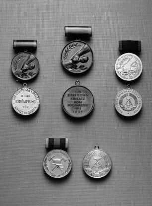 Drei Medaillen der Deutschen Demokratischen Republik. Oben: Medaille für selbstlosen Einsatz bei der Bekämpfung von Katastrophen (1957 gestiftet). Links: Erstes Modell (bronzefarben; 30 mm Durchmesser; 1957/1971 verliehen). Rechts: Zweites Modell (bronzefarben; 30 mm Durchmesser; 1972/1990 verliehen). Beide an blaubezogener Spange (11 x 24 mm). Jeweils Avers, darunter Revers. Oben, Mitte: Medaille für die Bekämpfung der Hochwasserkatastrophe im Juli 1954 (Bronze; 35 mm Durchmesser; 1954 gestiftet) an Spange (dunkelblau bezogen mit rotem Streifen; 12 x 27 mm. Avers, darunter Revers. Unten: Medaille für treue Dienste in der Freiwilligen Feuerwehr (1959 in drei Stufen gestiftet, ab 1978 vier Stufen). Medaille in Silber oder Gold (versilbert oder vergoldet; 31, 5 mm Durchmesser) an Spange (bezogen; 12 x 25 mm). Avers, daneben Revers
