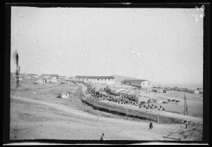 Nador (Marokko): "Kirchenparade zur Feldmesse" der Infanterie und Regimentsmusik [des spanischen 10. Kavallerie-Regiments "Alcántara" (?)] in hellgestreiften Uniformhosen, dunklen Uniformjacken, Tropenhelmen und Marschgepäck auf einer Ortsstraße mit Gebäuden am Straßenrand (Distanzaufnahme)