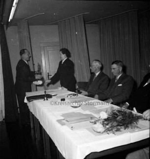 Keck, Erika: Kreispräsidentin: Kreisberufschule Bad Oldesloe: Kreistagssitzung am 18. Mai 1955: Verabschiedung und Überreichung eines Blumenstraußes: Präsidiumstisch mit dem neuen Kreispräsidenten Friedrich Hardt und den Stellvertretern Willy Esser und Paul Manthey: hinten Vorhänge