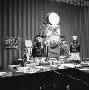 Oldesloer Spielmannszug e.V.: Jahresversammlung: Hotel Oldesloer Hof: leerer Vorstandstisch mit Platzkärtchen der Funktionsträger: hinten Ausstellung von Instrumenten und Standarten: daneben zwei Mädchen in Uniformen des Spielmannszugs, 15. Februar 1975