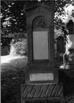 Grabstein 45 (Index: 100086687) Material: Sandstein mit zwei weißen Marmorplatten Zustand: Sandstein teilweise abgeblättert Maße HxBxT: 177x58x24 cm Beschreibung: ädikulaartige Stele mit Pilastern, getreppter Sockel, giebelartiger Abschluß mit Verdachung und Eckakroteren Symbolik: negatives Zahnfries, Mohnkapseln, Blüten, Ranken: Grabstein 45