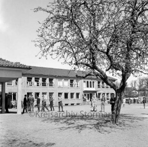 Volksschule am Schweinemarkt [später Emil-Nolde-Schule]: hinten Klassentrakt: links offene Pausenhalle: auf Schulhof Schüler: vorne Baum