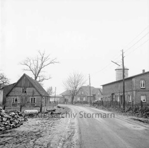 Dorfstraße mit Bauernhäusern und Silo: Ortsdurchfahrt: links Stapel mit Holzklötzen