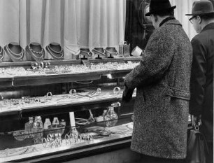 Männer stehen vor der Schaufensterscheibe eines Juweliergeschäftes und betrachten den in der Auslagen befindlichen Schmuck. Diese Art von Geschenken sind teuer