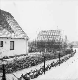 Neubausiedlung: links Einfamilienhaus mit Vorgarten, Hecke und Zaun: hinten rechts Rohbau mit Dachstuhl: Winter