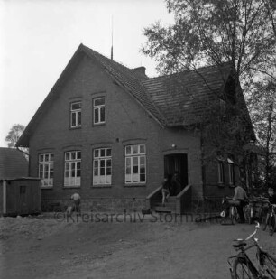 Volksschule: Schulkoppel: altes Schulgebäude: vorn Schulkinder: rechts Fahrradständer mit Fahrrädern: dahinter Bäume: links Baubude