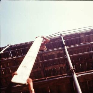 Zimmerer auf einem Gerüst beim vertikalen Weiterreichen einer Holzbohle