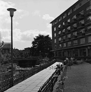 Grünanlage mit Fußweg, Jägerzaun und Straßenlampe: rechts Wohnblock mit Möbelhaus Bohl: links Trave: hinten links Brücke an Hagenstraße: dahinter Gebäude