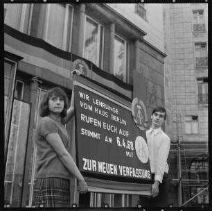 Volksentscheid zur neuen Verfassung der DDR am 6. April 1968, Bild 2: Junge Frau und junger Mann halten ein Schild, das für die Annahme der neuen Verfassung wirbt vor dem "Haus Berlin" am Strausberger Platz. SW-Foto, März 1968 © Kurt Schwarz.