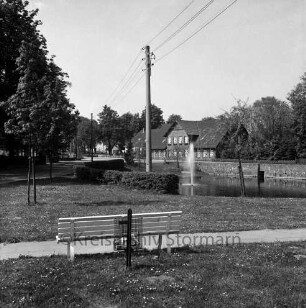 Dorfstraße: Dorfteich mit Springbrunnen: vorn Grünanlage mit Weg, Bank und Papierkorb: links Strommasten: hinten Häuser und Bäume