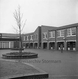 Bargteheide: Kreisgymnasium II [später Gymnasium Eckhorst]: Eckhorst: Außenaufnahme mit Uhr: davor Schulhof, Mann, Rundbeete mit Baum und Sitzfläche