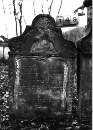 Grabstein 149 (Index: 99751970) Material: Sandstein Zustand: verwittert mit Textverlust, abgeblättert Maße HxBxT: 126x74x10 cm Beschreibung: Stele, in Voluten auslaufend. Rundbogenaufsatz, rechteck. Schriftfeld eingetieft Symbolik: Krone, Voluten, Ranken: Grabstein 149