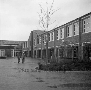 Bargteheide: Kreisgymnasium II [später Gymnasium Eckhorst]: Eckhorst: Außenaufnahme mit Schulhof: zwei Männer: vorne links Notausstieg aus Kellergeschoss, Tarnung als Brunnen