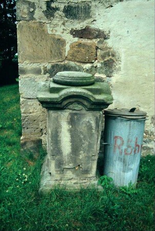 Grabstein am Eingang der Kirche; daneben Mülltonne