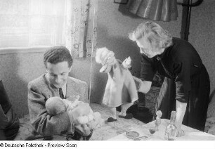 Mann mit einem Säugling im Arm, danebenstehend eine Frau mit Handpuppe
