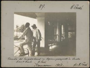 Selbstportrait mit Leutnant Frank im Regierungskrankenhaus von Douala : Seitliche Rückansicht von zwei Männern, am Geländer des Arkadengangs des sogenannten "Europäerhauses"stehend