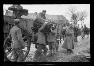 Nicht identifizierbare Ortschaft in Nordfrankreich oder Belgien: Soldaten mit Feldmützen neben einem hohen Pferdewagen auf einer morastigen Ortsstraße beim Ausladen