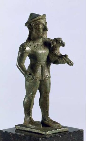 Archaische Votivstatuette eines bärtigen Mannes mit Chiton, Hut und Schnabelschuhen, einen Widder unter dem linken Arm tragend