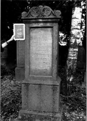 Grabstein 167 (Index: 100111340) Material: Sandstein Zustand: leicht verwittert Maße HxBxT: 197x60x46 cm Beschreibung: Stele, profilierter Sockel, Abdeckplatte mit Volutenaufsatz, rechteckige Schriftfelder eingetieft Symbolik: Blüten, Voluten: Grabstein 167