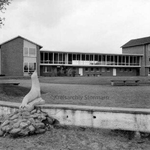 Realschule Harksheide: Fadens Tannen: Kunst im öffentlichen Raum: links Steinskulptur einer Robbe mit Steinhaufen auf kleiner Mauer: dahinter Schulhof mit Grünanlage, Sträuchern und Bank: hinten Schulgebäude