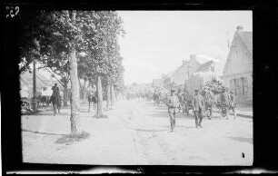 Origny (=Origny-Sainte-Benoite, Dep. Aisne, Frankreich): Soldatentrupp mit Feldmützen und umgehängten Gewehren und Pferdegespannen auf einer unbefestigten Ortsstraße, im Vordergrund 2 Offiziere in Gegenrichtung (Rückansicht, Distanzaufnahme)