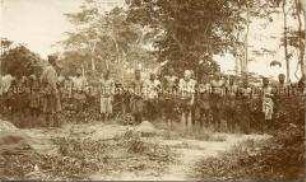 Angekettete Gefangene, darunter ein "Albino-Zauberer", bewacht von afrikanischen Soldaten der Schutztruppe