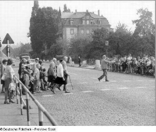 Ansammlung von Kindern und Erwachsenen zur Verkehrserziehung durch zwei Volkspolizisten: Geleiten von sehbehinderten Menschen auf einem Fußgängerüberweg