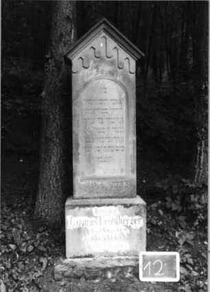 Grabstein 12 (Index: 100090870) Material: Sandstein Zustand: leicht verwittert Maße HxBxT: 157x42x18 cm Beschreibung: Stele, getreppt. Sockel, giebelart. Abschluß mit Verdach. , Schriftfeld m. Rundbogenabs Symbolik: gestelztes giebelartiges Fries: Grabstein 12
