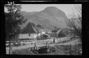 [Rucãr (?) (Rumänien)]: unbefestigte Ortsstraße über eine kleine Brücke, dahinter Wohnhäuser mit Mansardendach aus Schindeln, im Hintergrund ein Bergmassiv