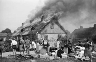 Brand: Meddelskamp: Reetdachbaracke als Flüchtlingsunterkunft: brennendes Dach: darüber Rauchschwaden: vorn auf Weg und Wiese Bewohner und Helfer sowie gerettetes Mobiliar: dahinter rechts und links Hecken und Gärten: hinten links Anbau: hinten rechts Reihenhaus