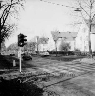 Sieker Landstraße: Fußgängerampel mit Zebrastreifen: links zwei Pkws: dahinter Mitte parkender Pkw: hinten rechts drei Wohnblocks mit Grünanlage, Hecke und Bäumen