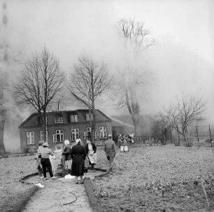 Brand: Wohn- und Wirtschaftsgebäude von Landwirt Leinius: Frauen mit geretteten Haushaltsgeräten im Garten: dahinter brennendes Wohnhaus hinter Baumreihe