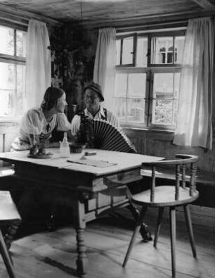 Bauernstube mit jungem Paar, an einem Eßtisch sitzend. Der Mann auf einem Knopfkasten spielend (vielleicht in Altstädten)