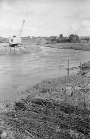 Trave: Flussregulierung: vorn Reisigbündel: links am Ufer Bagger, Loren und Baumaterial: hinten rechts und links des Ufers Wiesen und Büsche: dahinter Häuser von Sehmsdorf zwischen Bäumen