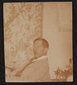Rudolf Borchardt sitzt nach links gewendet, Blick über die Schulter in die Kamera vor einem chinesischen Wandteppich