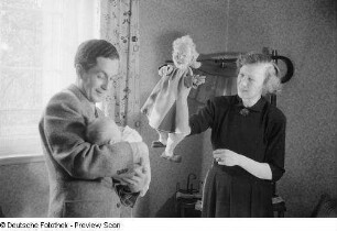 Mann mit einem Säugling im Arm, danebenstehend eine Frau mit einer Handpuppe