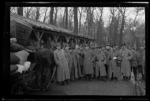 Nicht identifizierbare Ortschaft in Nordfrankreich oder Belgien: Gruppe von Offizieren mit Schirmmützen und Soldaten mit Feldmützen auf einer unbefestigten Straße neben einem lang gestreckten Holzschuppen am Rand eines Parks
