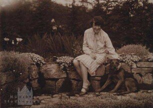 Auf einer Steinmauer im Garten sitzende Dame mit Schäferhund