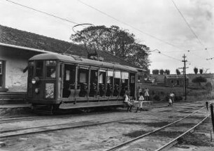 Straßenbahn : Eine Straßenbahn in einem Bahnhof. Ein Junge sitzt auf dem Trittbrett der Bahn, ein weiterer steht daneben. Im Hintergrund vier Mädchen