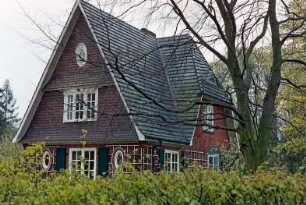 An den Stücken (7): Wohnhaus aus Backstein: Giebelseite, ovale Fenster, Rankgerüst, an Giebel Schindelverkleidung, vorgewölbtes Sprossenfenster: rechts an Traufseite Zwerchhaus, Sprossenfenster, Rankgerüst: im Vordergrund Hecke