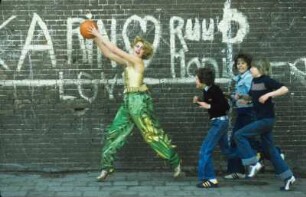 Aufnahme für die Reportagereihe „Außenseiter der Mode“: Studio Fong Leng, Amsterdam : Model in gelbem Top und grünen Pluderhosen nach Entwürfen von Fong Leng, mit einem Basketball und drei Jungs vor einer Graffitiwand