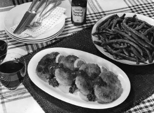 Krafts Rezeptaufnahmen. Platte mit Buletten mit Zitronenscheiben und Ketchup. Daneben eine Schale mit grünen Bohnen und eine Flasche Kraft's Tomatenketchup