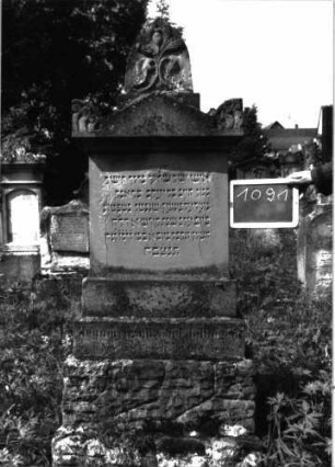 Grabstein 1091 (Index: 100104145) Material: Sandstein Zustand: teilweise angewittert Maße HxBxT: 178x53x20 cm Beschreibung: Stele auf getrepptem Sockel, Aufsatz mit Dreiecksabschluss und dahintergestellten spitzbogigen Giebel- sowie Eckakroterien, Giebelakroter mit Mohnkapseln, Eckakroterien mit Voluten, unterster Sockel felsartig gearbeitet, Stele wirkt unproportioniert;Mittelteil ist zu niedrig im Verhältnis zum Sockel Symbolik: Mohnkapseln, Voluten: Grabstein 1091